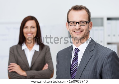 Handsome confident businessman in glasses standing facing the camera with a friendly smile with an attractive female coworker in the background