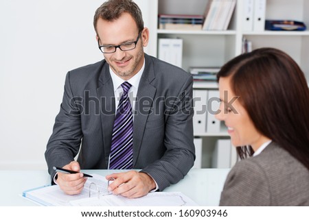 Business partners at work in the office with focus over the shoulder of a woman to a smiling confident businessman in glasses working on paperwork