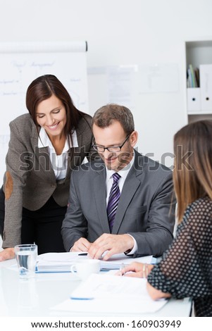 Team of businesspeople in a meeting with focus to a smiling woman standing over the shoulder of a man in glasses as they discuss paperwork spread out on the desk