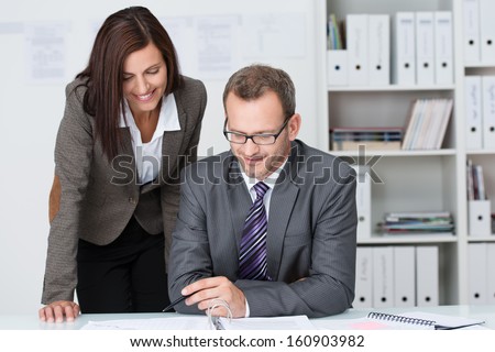 Businessman seated at his desk working with his secretary as the two look at a document together with his secretary, an attractive young woman, standing leaning over his shoulder