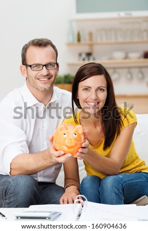 Enthusiastic attractive young couple planning for the future sit side by side on a sofa in the living room holding a piggy bank with an open file and calculator in front of them