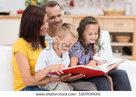 Happy Family Reading A Book Together With Attractive Young Parents Grouped With Their Two Small Children On A Couch In The Living Room