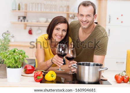 Cheerful young couple cooking a meal leaning side by side on the kitchen counter surrounded by fresh ingredients and each holding a glass of red wine