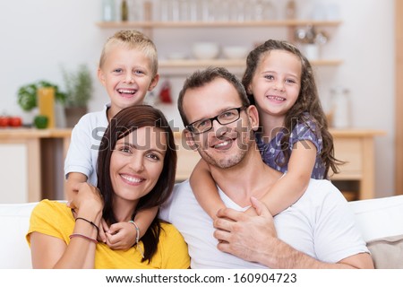 Laughing confident young family with the two small children standing with their arms around their parents necks as they relax together on a sofa in the house