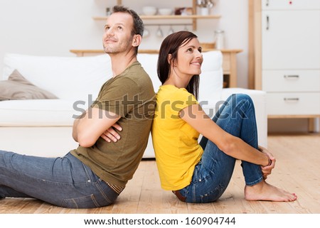Young Couple Relaxing On The Floor At Home Sitting Barefoot Back To Back Each Looking Up Into The Air With A Thoughtful Smile As They Plan Their Future Together