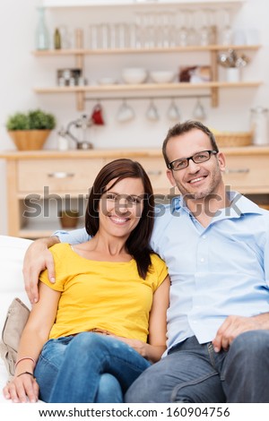 Affectionate Couple Relaxing Together On A Sofa In The Living Room Sitting Side By Side Giving The Camera Charming Cheerful Smiles