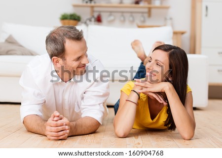 Loving Young Couple Enjoying A Relaxing Day Lying Facing Each Other And Smiling On The Wooden Floor In The Living Room