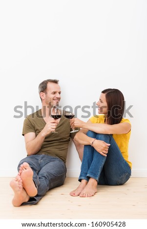 Happy young barefoot couple sitting on the wooden floor in their living room smiling and toasting each other with a glass of red wine celebrating moving to a new house