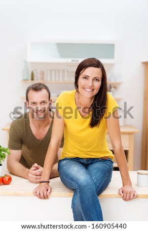 Beautiful young woman sitting on the counter top in the kitchen smiling at the camera with her husband leaning on the counter behind her