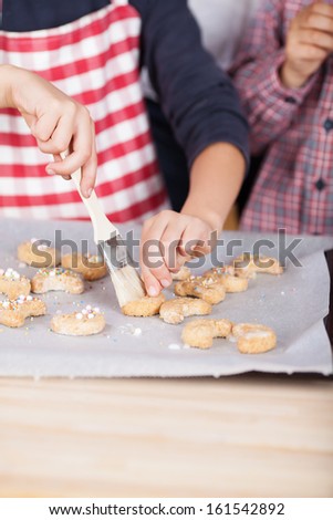 Close up view of the hands of a young child in a red and white striped apron decorating cookies with a pastry brush and sugar pearls