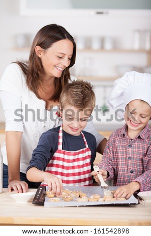 Two happy young brothers, one in a chefs toque and one in a colorful red and white apron, baking cookies in the kitchen watched over by their attractive mother