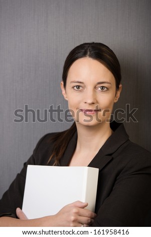 Young attractive female business executive clutching a file to her chest standing looking at the camera with a smile on a grey background