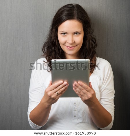 Happy young woman with a lovely smile standing reading information on her handheld tablet computer against a grey studio background