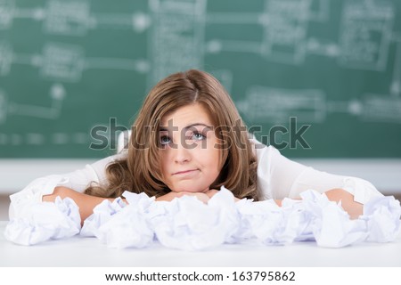 Beautiful young teenage college student at wits end and unable to find a solution or fresh ideas leaning on the desk surrounded by crumpled paper making a whimsical expression