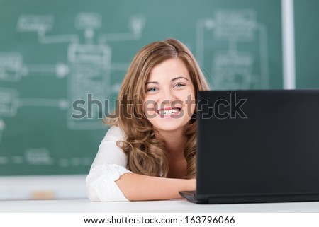 Smiling teenage girl in a college classroom sitting in front of the black board looking around her laptop screen at the camera