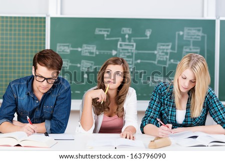 Thoughtful attractive young college student sitting at a desk in the classroom with two teenage classmates staring thoughtfully up into the air as she seeks an answer and solution