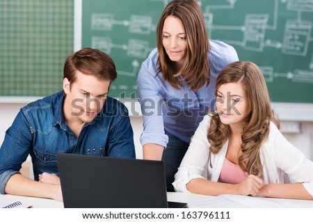 Teacher working with two teenage students in class bending over between a young teenage boy and girl as they read the laptop computer screen together