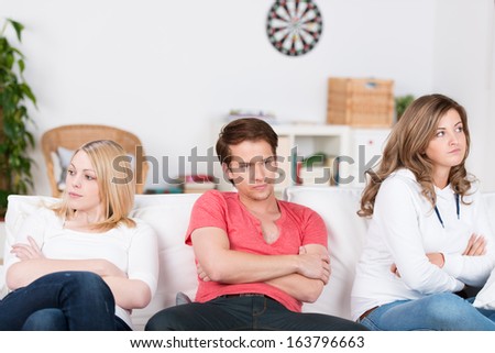 Three Young Friends After An Argument Sitting In A Row On A Sofa In The Living Room With Folded Arms And Angry Expressions Ignoring One Another