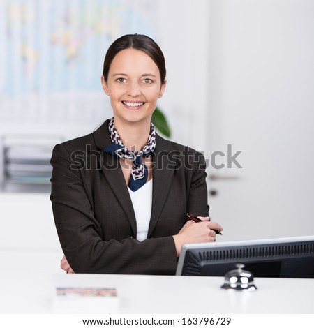 Welcoming confident friendly receptionist standing behind a hotel reception desk with her arms folded giving the camera a beautiful warm smile