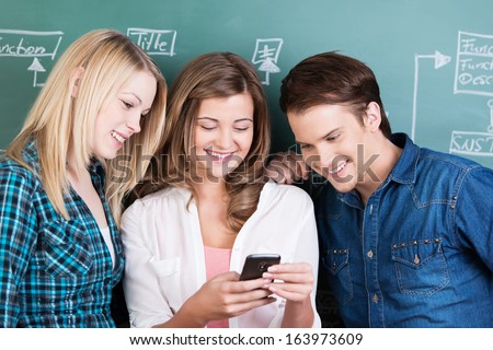 Group of three young teenagers smiling as they read a text message on the mobile of a beautiful girl in the classroom during a break in classes