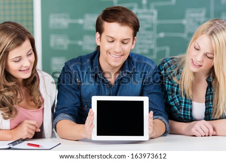 Group of three attractive young teenage students displaying a blank tablet-pc with the screen to the viewer as they sit in front of a class blackboard
