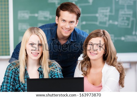 Three confident happy teenage college students posing together behind a laptop computer smiling over the top at the camera with a blackboard background