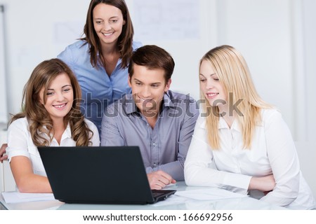 Three happy smiling young teenage college students working together on a laptop watched over by their smiling female teacher