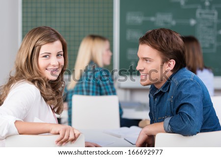 Young teenage girl and boy in class at college sitting sharing a desk and turning to look back over their chairs at the camera with classmates and the blackboard behind