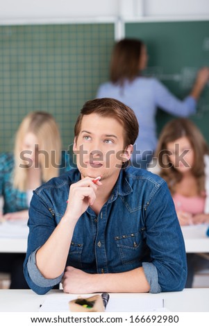 Young man in class trying to find a solution sitting at his desk in college with his hand to his chin staring upwards with a serious expression
