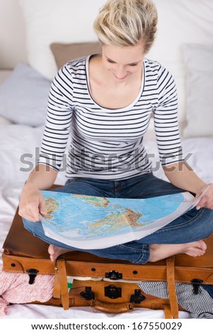 Woman looking for exotic travel destinations sitting cross legged on her suitcase on her bed reading a world map