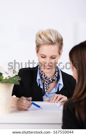 Beautiful receptionist assisting a client to check in at a hotel reception desk pointing to documentation that must be completed