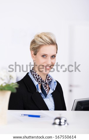 Beautiful elegant hotel young blond receptionist working at her computer at the counter looking up with a warm welcoming smile