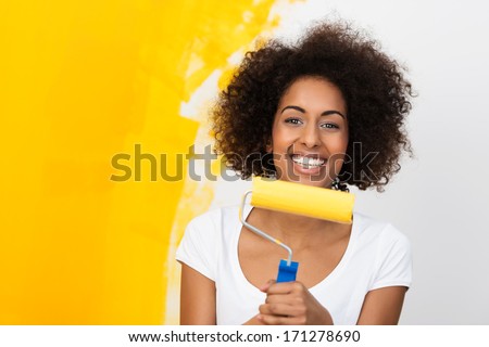 Smiling African American woman redecorating her house holding a paint roller covered in orange paint which she has been applying to the wall behind her Smiling African American woman redecorating her house holding a paint roller covered in orange paint which she has been applying to the wall behind her