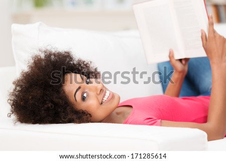 Happy relaxed young African American woman looking at camera while laying on her back on a white sofa, holding an open book