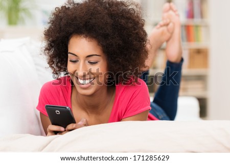 Beautiful African American woman with a frizzy afro hairstyle texting on her mobile while lying on her stomach on a sofa