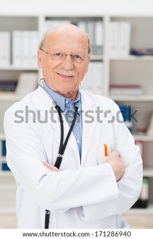 Friendly elderly male doctor with a stethoscope around his neck standing with folded arms in his office or surgery smiling at the camera