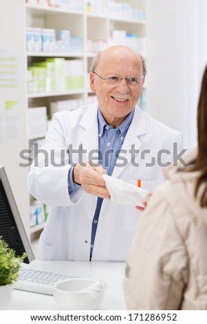 Friendly elderly male pharmacist standing behind the counter in the pharmacy dispensing prescription medicine to a patient with a smile