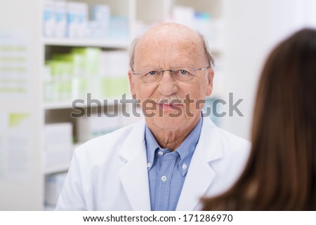 Elderly male pharmacist serving a patient in the pharmacy, view over the womans shoulder to focus on his face