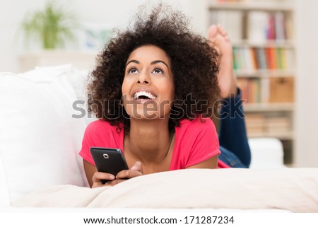 Happy African American Woman Thinking What To Say In A Text Message Lying On Her Stomach On A Sofa Looking Into The Air For Inspiration While Holding Her Mobile Phone