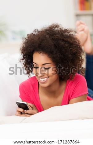 Smiling Beautiful Young African American Woman With An Afro Hairstyle Lying On A Sofa Reading An Sms On Her Mobile Phone