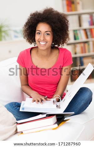 Cheerful African American female student, wearing casual clothes, sitting in the lotus position on the sofa, while holding an open folder and having different books in front of her