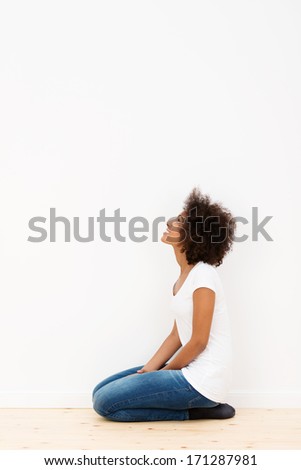 African American woman kneeling on the floor looking at a white wall in her new house as she visualizes her new color scheme and decor, with copyspace