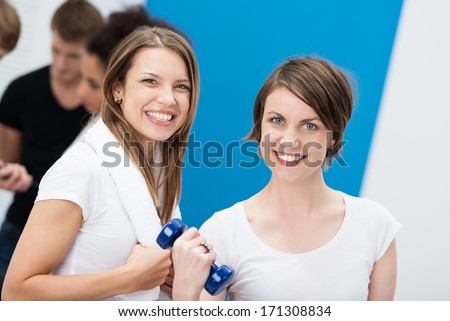 Two vivacious young women at the gym smiling at the camera as one works out with dumbbells to tone her muscles