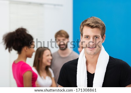 Smiling handsome young man at the gym posing in front of a group of his friends with a towel around his neck looking at the camera