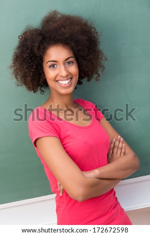 happy smiling African American woman with a frizzy afro hairstyle and a beautiful friendly smile standing with folded arms against a green blackboard