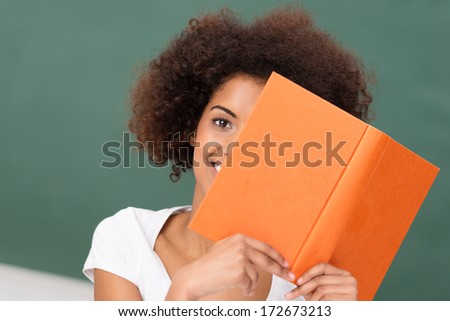 African American woman with a frizzy afro hairstyle reading an orange hardcover book peeping around the edge with one eye at the camera