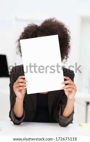 Businesswoman with a bushy afro hairstyle hiding behind a blank sheet of paper covering her face as she sits at her desk