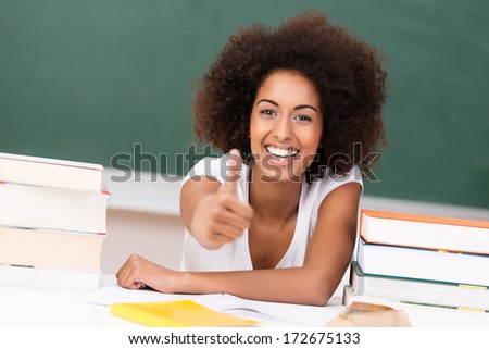 Laughing vivacious young African American woman student giving a thumbs up of approval and success as she sits at her desk with her text books