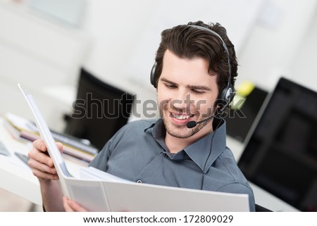 Call Centre Operator Or Client Services Businessman Wearing A Headset Looking Up Information In A File In Order To Be Of Assistance To The Client