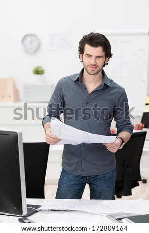 Businessman dealing with paperwork in the office standing behind his desk with an open spreadsheet in his hand smiling at the camera